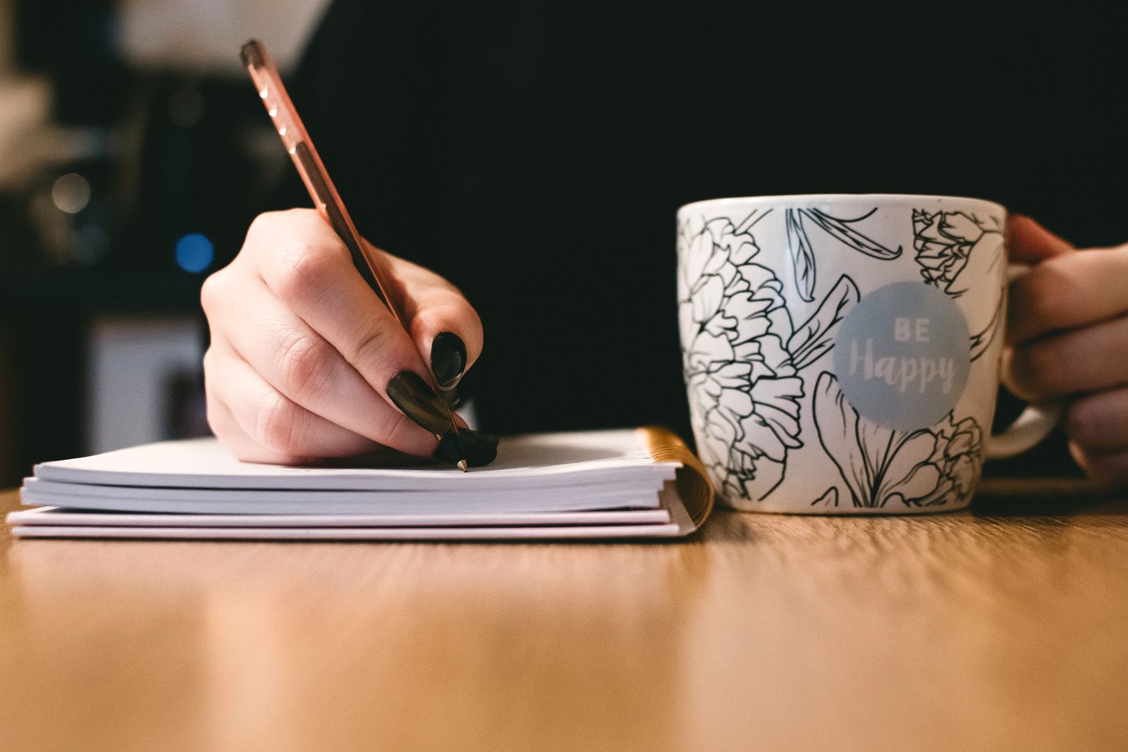 A hand writing in a notebook with a pen, a cup of coffee beside it