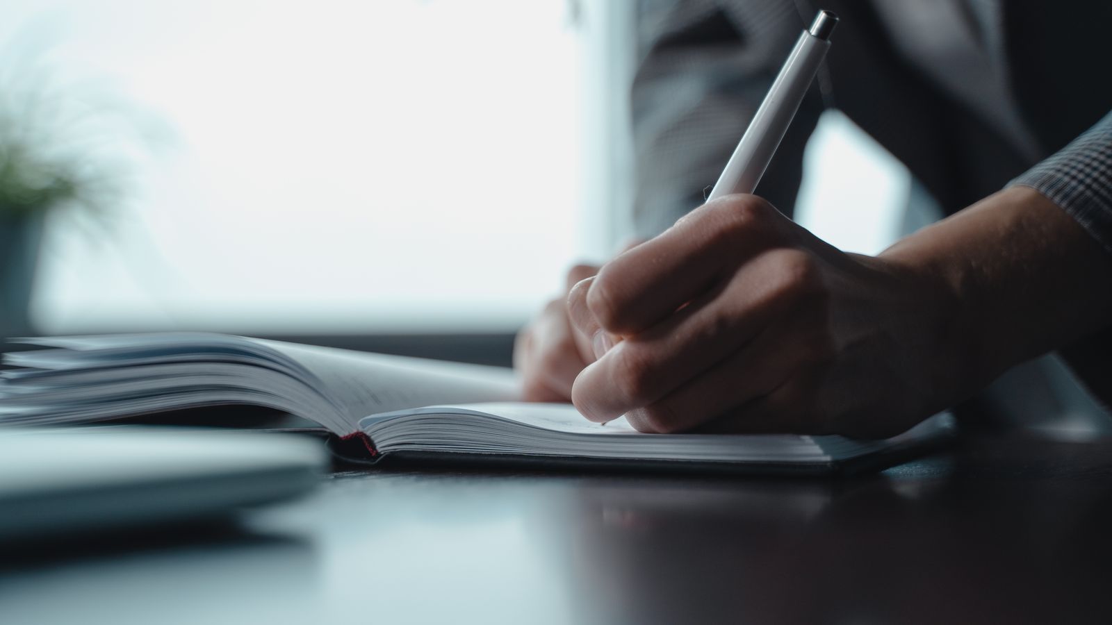 A hand writing in an open notebook beside a window