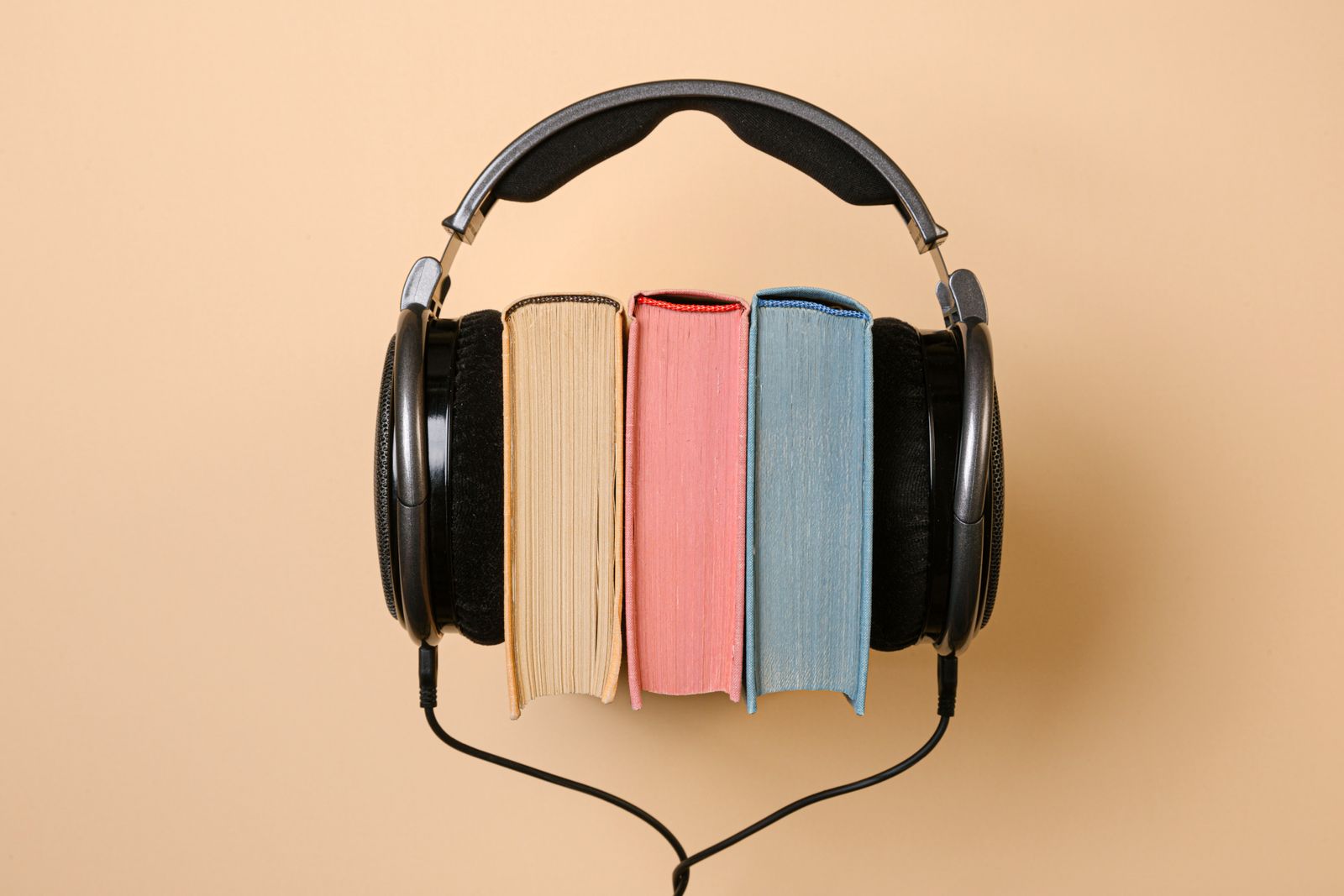 A large pair of headphones wrapped around three books in beige, pink and light blue against a warm background