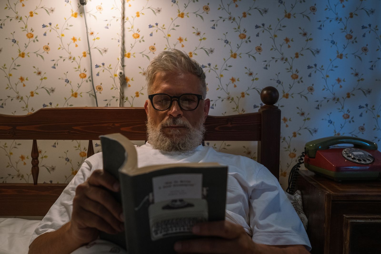A man with glasses and a beard reads a paperback in bed at night, floral wallpaper and a red telephone behind him