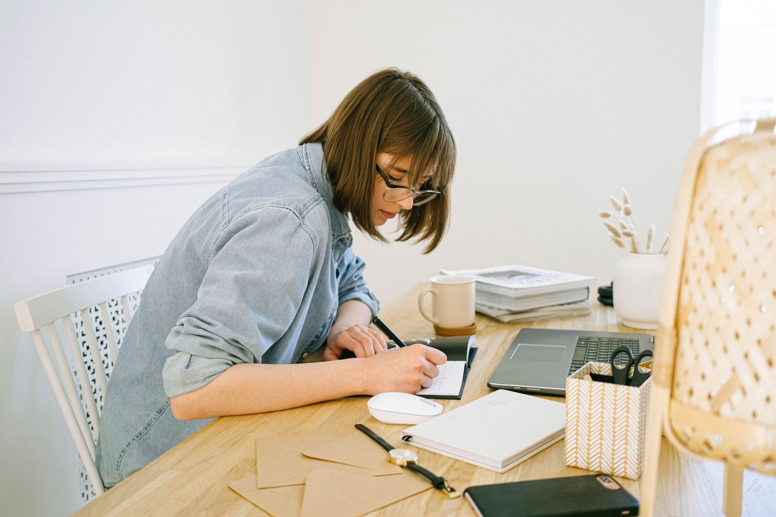 A woman with glasses writes in a notebook at a bright wooden desk, an open laptop next to her