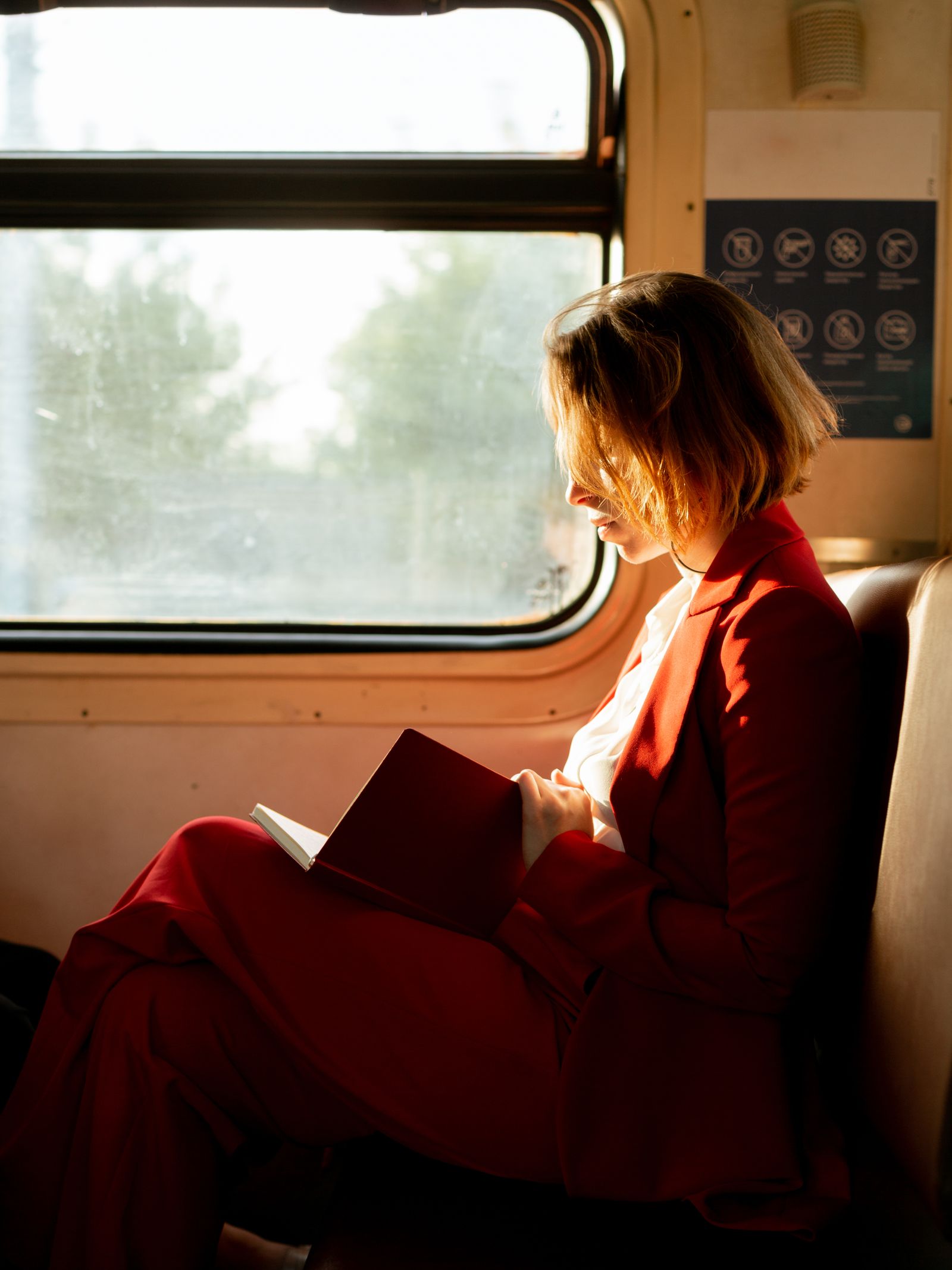 A woman in a red suit reads by the window of a train, sunlight streaming in